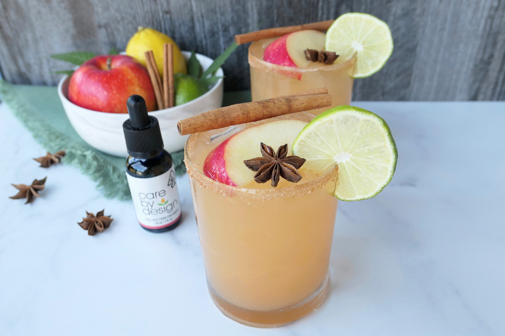 Photo of Spiced Apple CBD Margarita on a table beside a bowl of fruit and a Care By Design tincture bottle